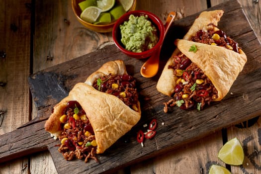 From above of traditional Mexican enchiladas prepared with rolled corn tortilla and stuffed with spicy meat and bean with chili pepper served with guacamole sauce and fresh lime on rustic wooden table