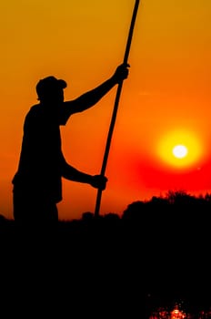 A skilled poler pushes his dugout boat forward in the Okavango Delta, while standing upright in the boat, and seen as a silhouette, arms stretched out, while holding the pole in both hands, behind him the yellow glow of the sun setting in a beautiful red orange sky.