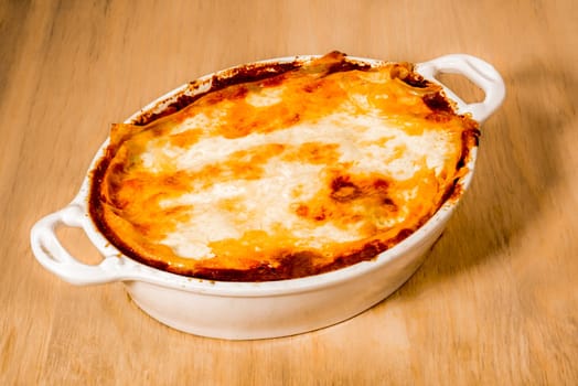 A delicious looking dish of lasagna with a golden yellow melted layer of cheese on top, inside a white ceramic dish, placed on a wooden surface.