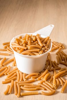 A standard cup size measuring cup of white plastic sits on a wooden surface, filled to the brim with wholewheat macaroni, while much more macaroni lies spilled around the cup.