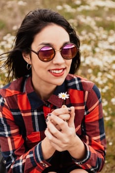 Beautiful woman with sunglasses sitting in the middle of a flowery meadow smelling a daisy