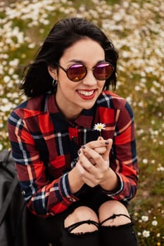 Beautiful woman with sunglasses sitting in the middle of a flowery meadow smelling a daisy