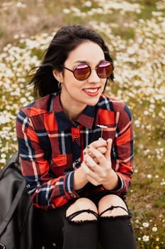 Beautiful woman with sunglasses sitting in the middle of a flowery meadow smelling a daisy