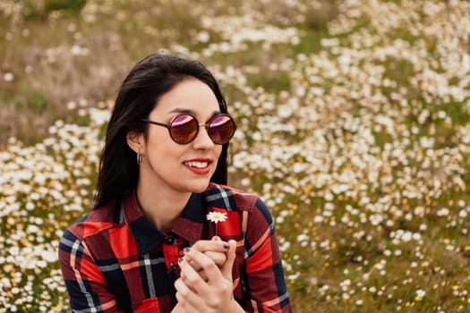 Beautiful woman with sunglasses sitting in the middle of a flowery meadow smelling a daisy