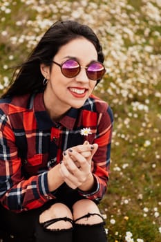 Beautiful woman with sunglasses sitting in the middle of a flowery meadow smelling a daisy