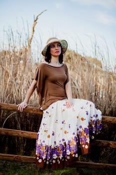 Stylish woman with long skirt sitting on the top of a wooden fence in the field