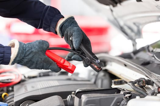 Human trying to start engine with jumper cables in close up. Emergency vehicle start-up. Hands in winter gloves holding black and red tool over open hood of a car.