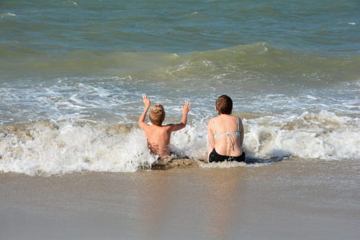 Two children sit full of joy on the sandy beach in the middle of the waves by the sea, on the North Sea coast in the Netherlands, Zeeland