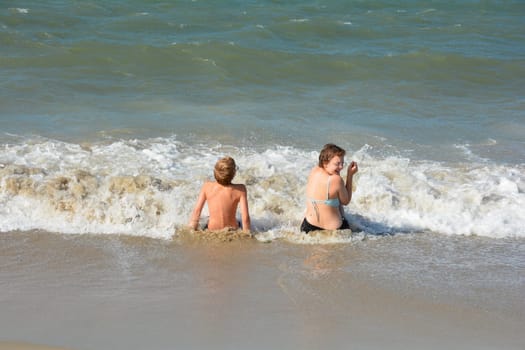 Two children sit full of joy on the sandy beach in the middle of the waves by the sea, on the North Sea coast in the Netherlands, Zeeland