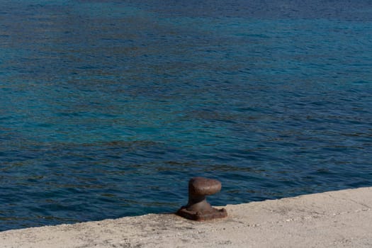 A rusty bollard on a quay wall with a rusty chain and in the background the water in the harbor on the Mediterranean Sea.