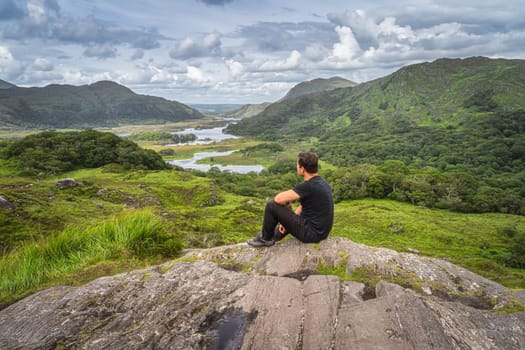 Middle age man sitting on a rock and admiring beautiful Ladies View, one of iconic Irish viewpoints, Lakes of Killarney, Rink of Kerry, Ireland