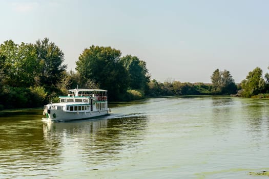 view of touristic passenger vessel cruising on Mincio river, shot in bright autumn sun light near Mantua, Lombardy, Italy.