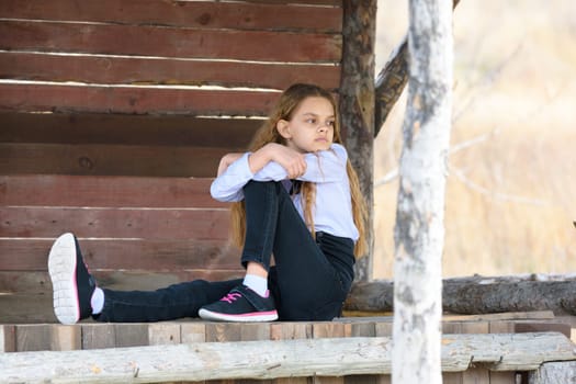 A girl sits on a wooden table against the background of a wall of boards and looks into the distance