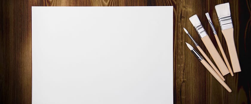 A clean white sheet and brushes on a wood textured background with space to copy. Layout, mockup space for text and drawing.