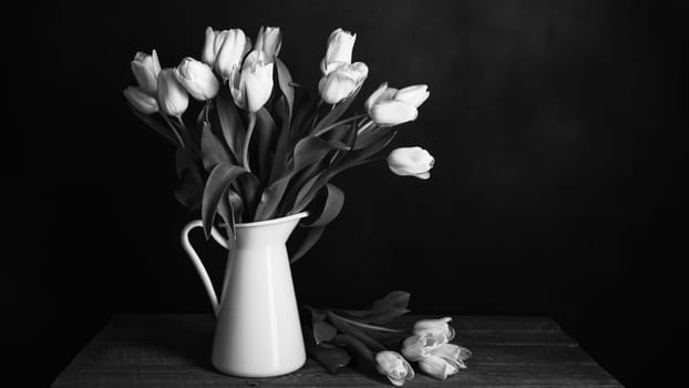 Tulips in a jug. Classic still life with a bouquet of tulip flowers in a vintage white jug on a dark background and an old wooden table. The style is black and white. Art photography.