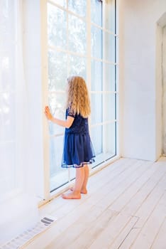 Cute girl in casual clothes standing near the window and watching out in light interior