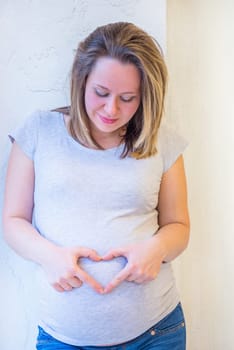 Beautiful pregnant woman standing near window at home in light interior and showing heart symbol from fingers