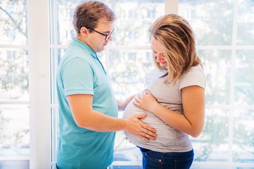 Beautiful pregnant woman and man standing near the window and holding belly