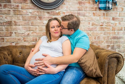 A man kissing his pregnant woman. They are sitting near a brick wall