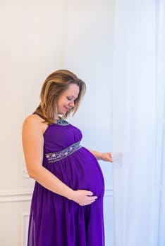 Beautiful pregnant woman in ultra violet dress standing near window in light interior