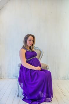 Beautiful pregnant woman in ultra violet dress sitting on the armchair in light interior