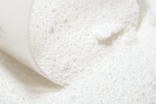 Macro image of clear plastic scoop measuring out white laundry detergent powder. Domestic cleaning chemical for clothing and fabrics.
