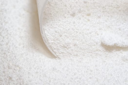Pure white laundry detergent powder, clumped together and being picked up by a clear plastic measuring scoop. Bright image and super macro perspective
