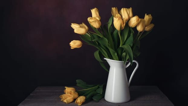Tulips in a jug. Classic still life with a bouquet of yellow tulip flowers in a vintage white jug on a dark background and an old wooden table. Art photography.