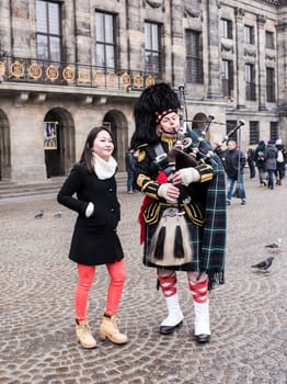 Amsterdam,Netherlands-December 30,2016:Bagpiper with his bagpipe in the square. Woman photographed with him. Nearby are walking tourists