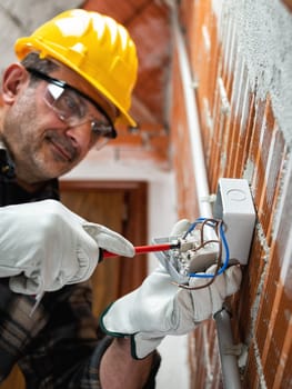 Electrician worker with a screwdriver fixes the cable in the switch of an electrical system  wear helmet, gloves and goggles. Workplace safety.