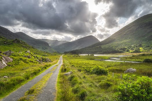 Straight country road leading trough Black Valley, dramatic sky. Landscape with lake, river and MacGillycuddys Reeks mountains, Ring of Kerry, Ireland