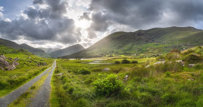 Straight country road leading trough Black Valley, dramatic sky. Landscape with lake, river and MacGillycuddys Reeks mountains, Ring of Kerry, Ireland