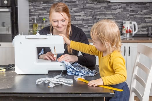 Smiling young mother is sewing and her son is holding sewing machine. Horizontally.