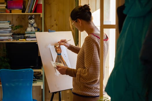 The artist paints a drawing of a cat with acrylic paints on an easel, in a small workshop by the window