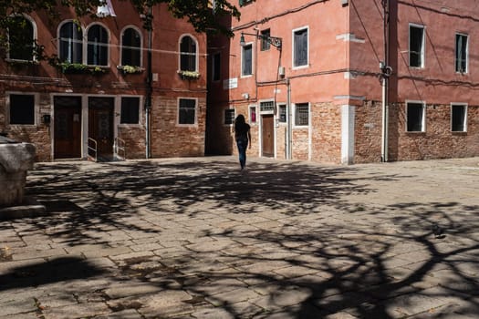 Bright sunlight with the shadows of the trees on a small square surrounded by old Venetian buildings of intense red color. A young girl walks alone through a "Campiello"", a narrow square surrounded by low buildings that leads to the ""Calli"" or Venetian alleys. One of the many micro neighborhoods of Venice (Italy) where in ancient times a large part of social life took place."