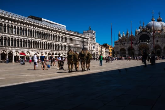 Rear view of some soldiers of the Italian army crossing Saint Mark's Square in Venice. They walk in the direction of the north side of the square, among tourists enjoying a sunny day. Frontally the complex of the Procuratie Vecchie with the portico of 50 arches. The square is dominated at its eastern end by St. Mark's Basilica..Perspective view with large shaded area.
