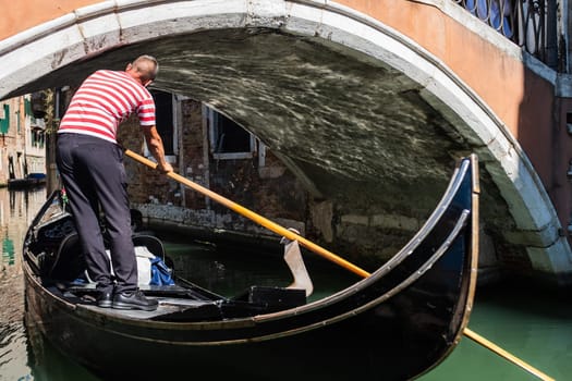 A gondolier lowers his head to pass under the bridge in a narrow canal in Venice (Italy).He is standing on the gondola and wearing the traditional clothing. The gondolier maneuvers skillfully through the narrow canals of the lagoon city.
