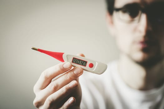 Man holds a red fever thermometer with high temperature in his hand, blurry face