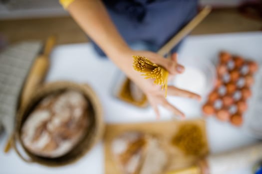 Female hand holding uncooked spaghetti above blurry ingredients on white table. Blurry eggs, pasta, flour, and bread on table from above. Pasta preparation