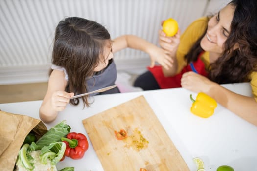 Happy little girl and her mother slicing lemons and tomatoes on cutting board. Smiling woman and young child at white talbe holding lemons. Daughter-mother cooking together