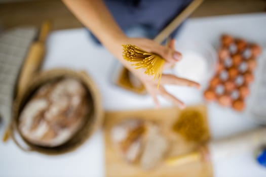 Female hand holding uncooked spaghetti above blurry ingredients on white table. Blurry eggs, pasta, flour, and bread on table from above. Pasta preparation