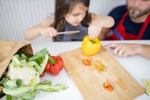 Little girl and her father slicing yellow bell pepper on cutting board. Man helping young child cutting vegetables on wooden board. Daughter-father cooking together