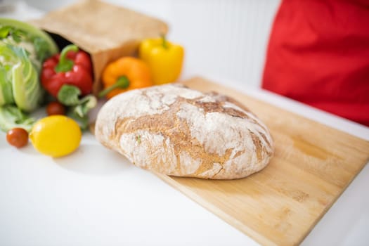 Bread on cutting board above white table and next to paper bag of vegetables. Sourdough bread alongside bell peppers, cauliflower, and lemons. Healthy meal preparation