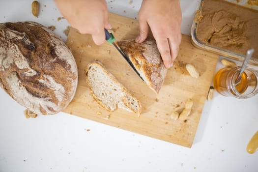 Female hands slicing bread on wooden cutting board from above. Woman cutting bread next to peanut butter and a honey jar. Healthy meal preparation
