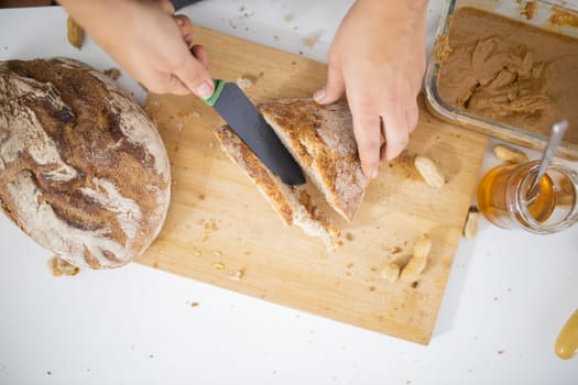 Female hands slicing bread on wooden cutting board from above. Woman cutting bread next to peanut butter and a honey jar. Healthy meal preparation