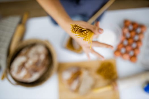 Female hand holding uncooked spaghetti above blurry ingredients on white table. Blurry eggs, pasta, flour, and bread on table from above. Pasta preparation
