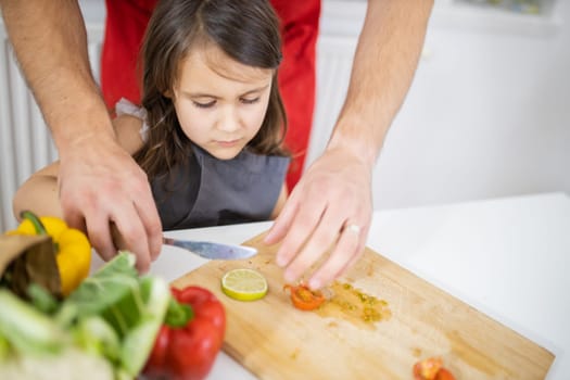 Little girl and her father slicing lemons, tomatoes, and bell peppers on cutting board. Male hands and young child cutting vegetables on wooden board. Daughter-father cooking together