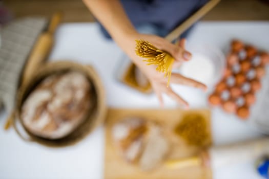 Female hand holding uncooked spaghetti above blurry ingredients on white table. Blurry eggs, pasta, flour, and bread on table from above. Pasta preparation
