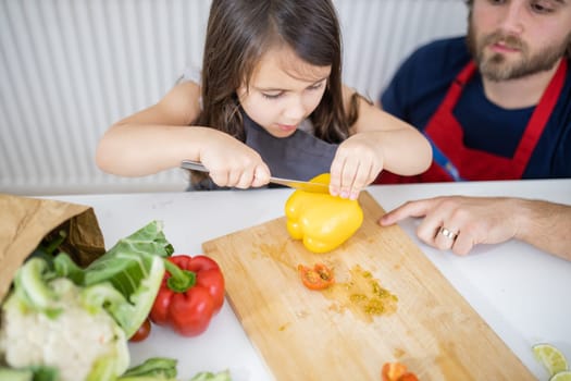 Little girl and her father slicing yellow bell pepper on cutting board. Man helping young child cutting vegetables on wooden board. Daughter-father cooking together