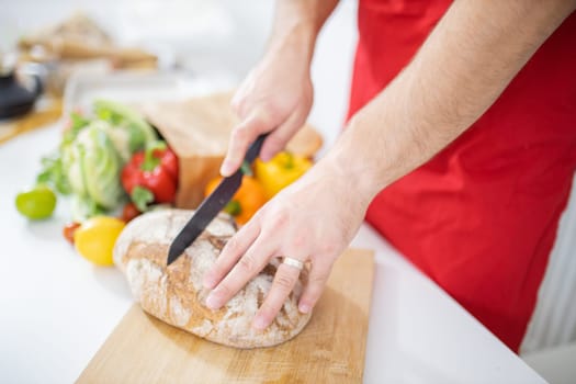 Male hands slicing bread on wooden cutting board above white table. Man cutting bread alongside bell peppers and cauliflower. Healthy meal preparation